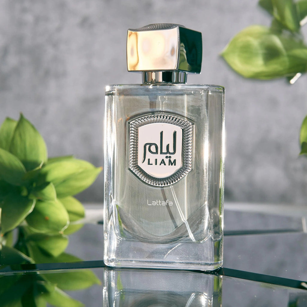 Clear glass perfume bottle with a silver cap on a reflective surface, surrounded by green plants.